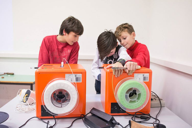 Three boys in a classroom interacting with two orange 3D printers, which have spools of white and green filament, suggesting a hands-on learning activity.