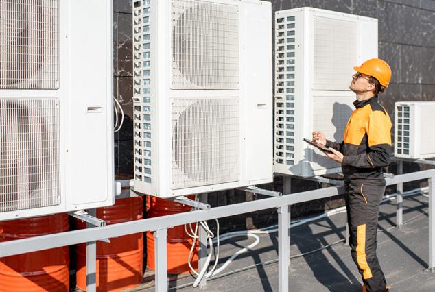 A technician in an orange hard hat inspecting multiple outdoor air conditioning units on a rooftop with a clipboard in hand.