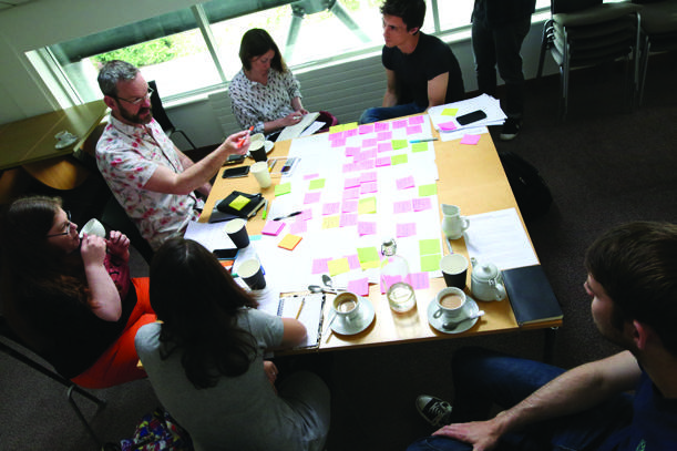 Overhead view of people collaborating around a table covered with pink and green sticky notes, coffee cups, and documents, indicating a brainstorming session.