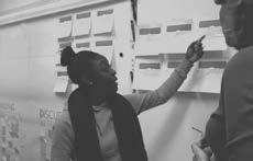 Two people in discussion, one pointing at sticky notes on a wall, likely engaged in a brainstorming or project planning session.