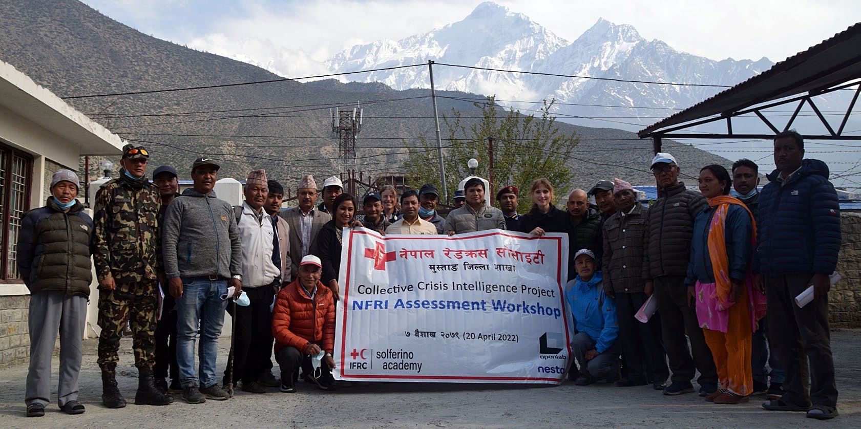 A group of people holding a banner for an "NFRI Assessment Workshop" outdoors in front of snowy mountains and a building.