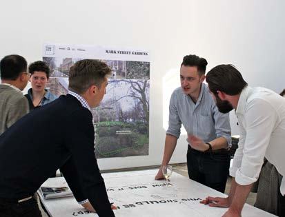 Group of people discussing urban planning in front of a poster for "MARK STREET GARDENS" project, with a large drawing on a table.