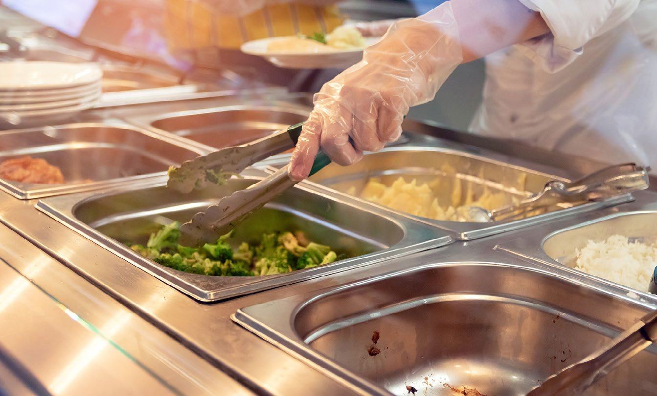 Gloved hand serving broccoli from a cafeteria buffet line with various hot food options.