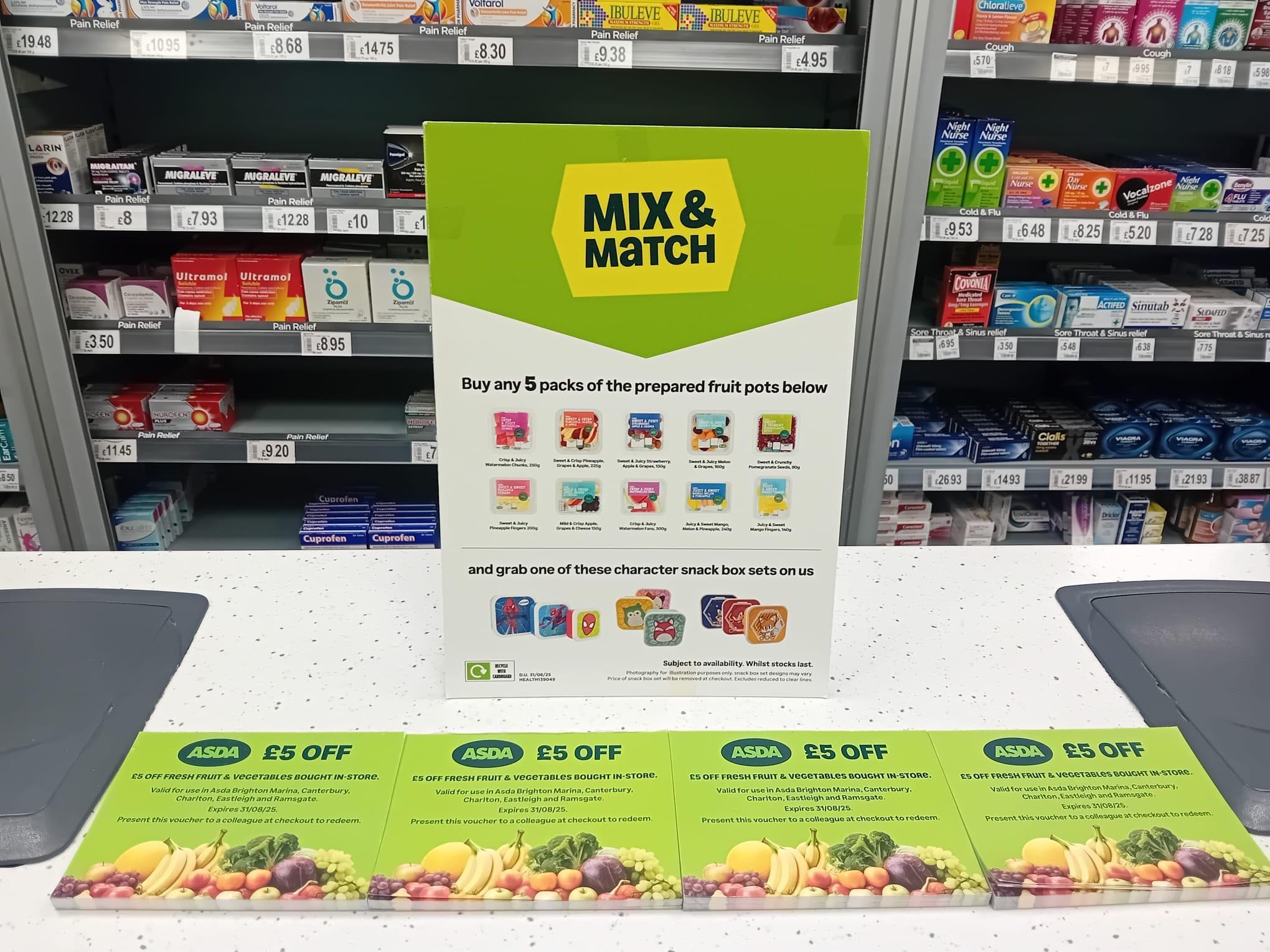 Retail display featuring a "Mix & Match" offer sign for fruit pots and snack boxes, alongside shelves of medications. In the foreground are ASDA vouchers for £5 off fresh fruit & vegetables, specifying store locations and expiry.