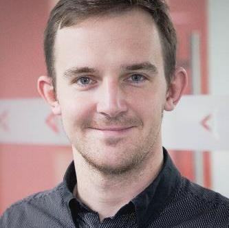 Headshot of a smiling man with short brown hair and beard, wearing a dark collared shirt.