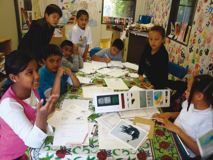 Diverse children sit around a table covered in papers and crafts, engaged in a learning activity, with one girl speaking.