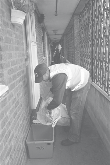 A person in a high-visibility vest and cap places items into a grey bin labeled '24/G' on an outdoor walkway.