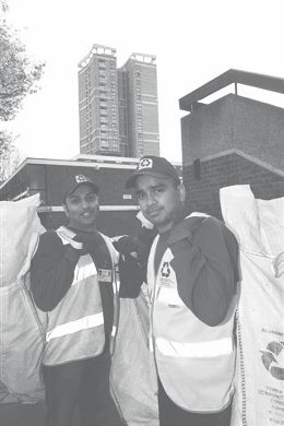 Two waste management or recycling workers in reflective vests and caps standing next to large collection bags, with buildings in the background.