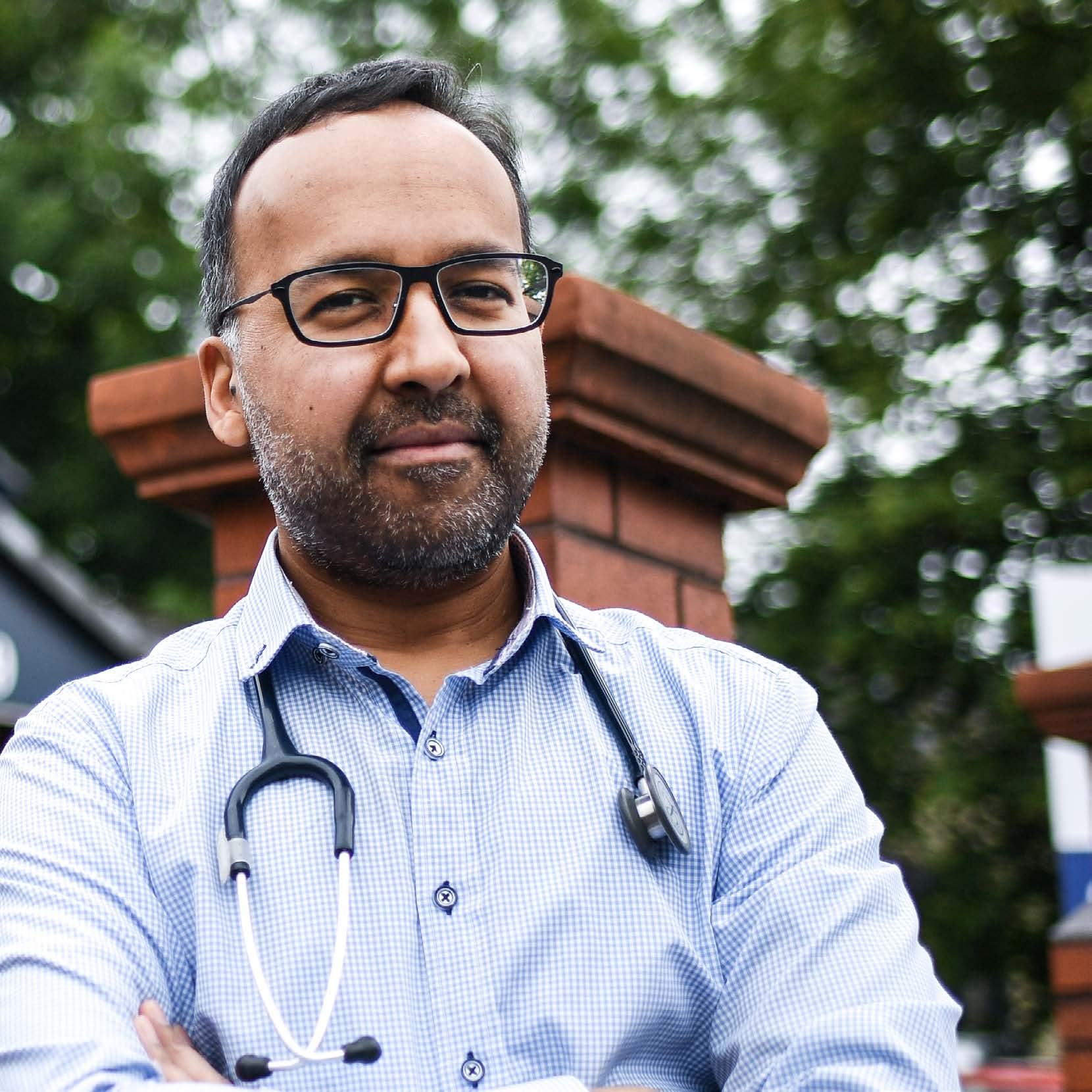 Headshot of a male doctor with a beard and glasses, wearing a light blue checkered shirt and stethoscope, looking at the camera.
