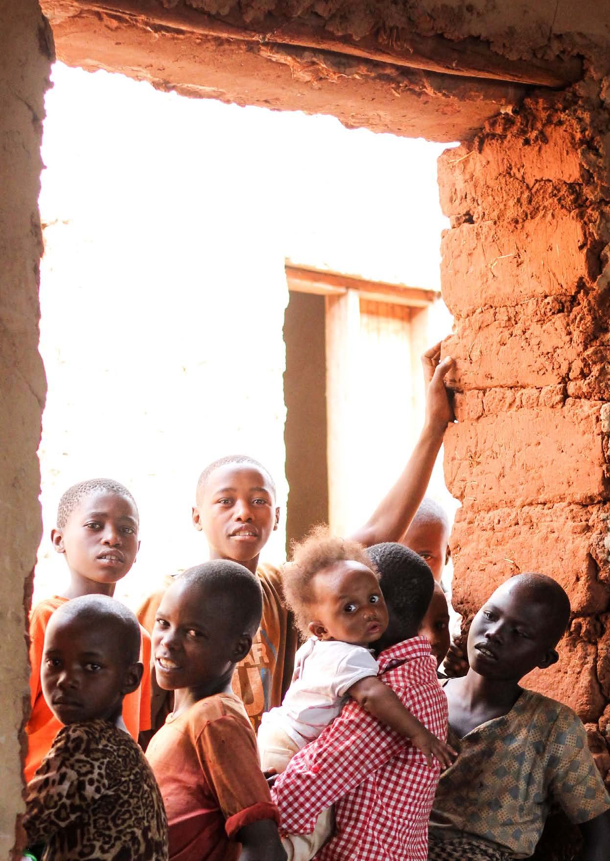 Group of children, including an infant, inside or near a doorway of a mud-built structure, with some looking towards the camera.