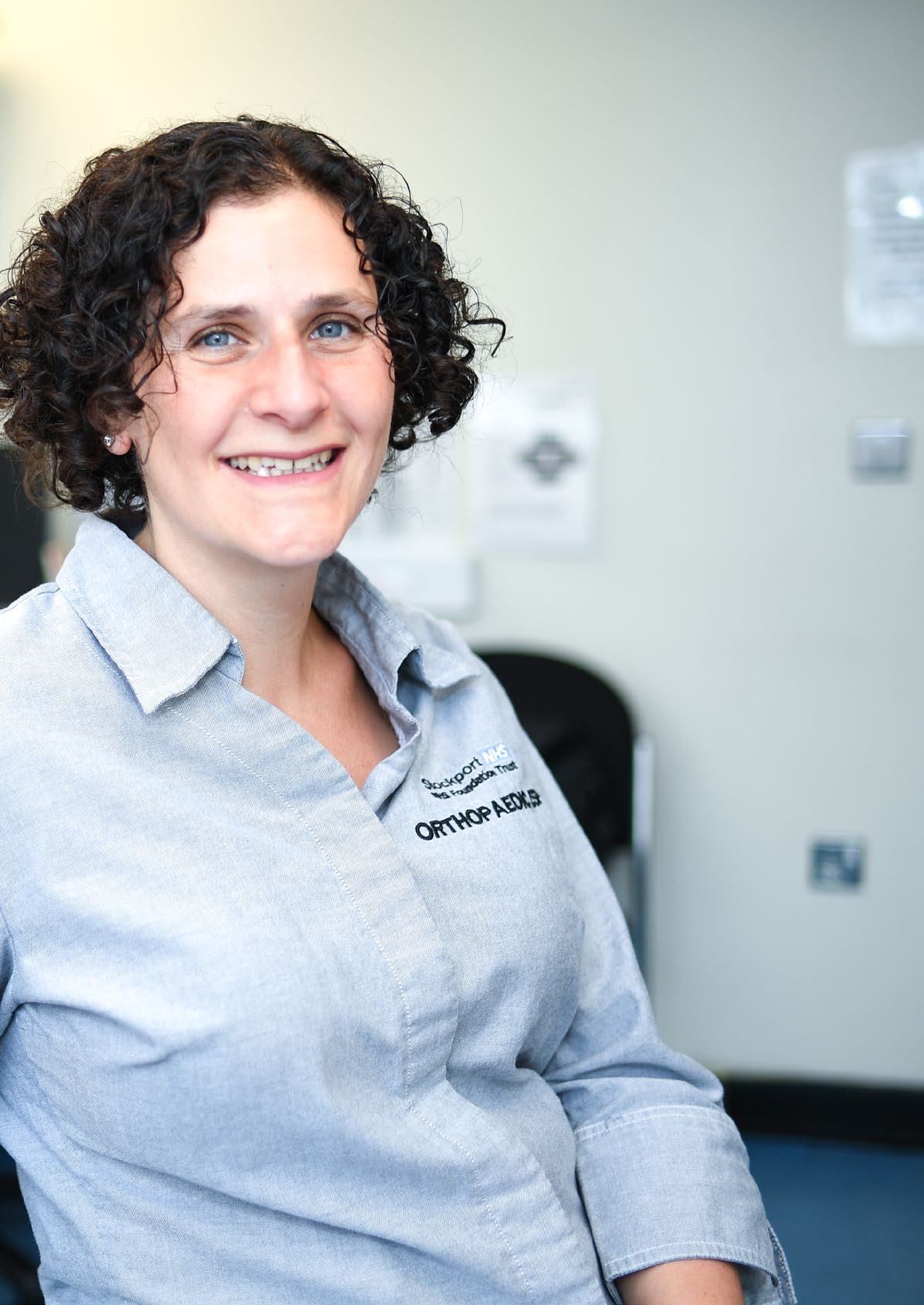 A smiling woman with curly dark hair wearing a shirt embroidered with "Stockport NHS Foundation Trust Orthopaedics".