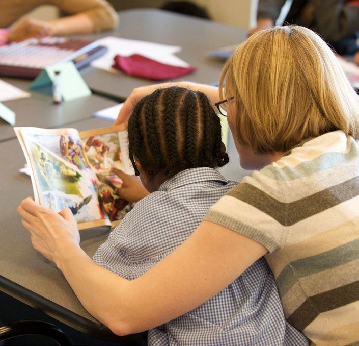 An adult with blonde hair reads a book with a young child with braided hair, both looking at the pages together.