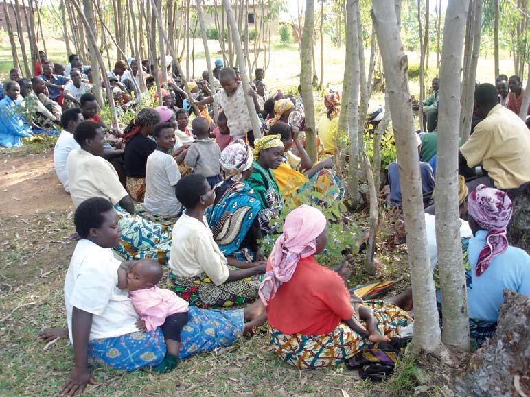 A large group of people, mostly women and children, are sitting on the ground outdoors amongst young trees in a community gathering.