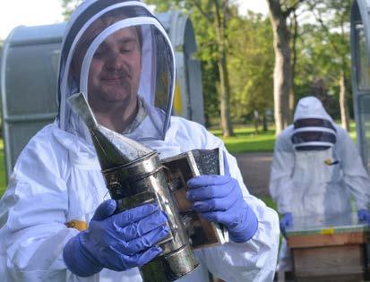 A person in a beekeeper suit holding a smoker, with another beekeeper visible in the background tending to a beehive.