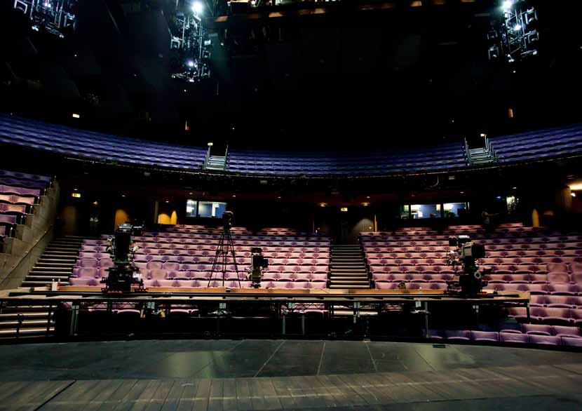 An empty theater viewed from the stage, showing rows of purple upholstered seats, a stage platform with equipment, and overhead lighting rigs.