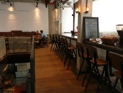 Interior view of a cafe or common area with a long bar counter, stools, a chalkboard menu, and people sitting at tables in the background.