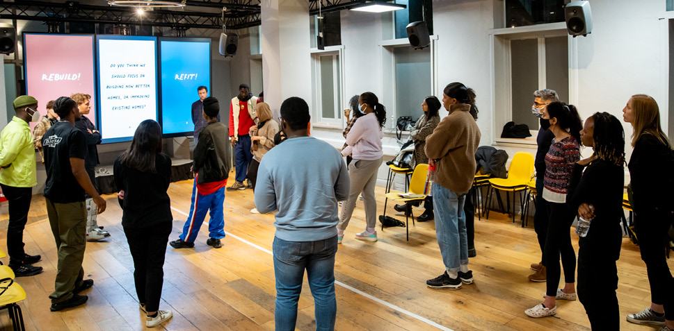 A group of people standing in a modern room, engaging in a discussion with large screens displaying text about housing and rebuilding.