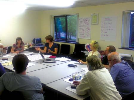 A group of people around a conference table in a meeting room, with notes on the wall and some participants engaged in discussion.