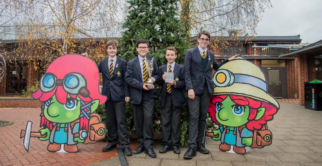 Four schoolboys in uniform, holding trophies, stand outdoors with two cartoon miner character cutouts, celebrating an achievement.