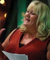 Woman with blonde hair speaking into a microphone, holding papers, against a green-lit background.