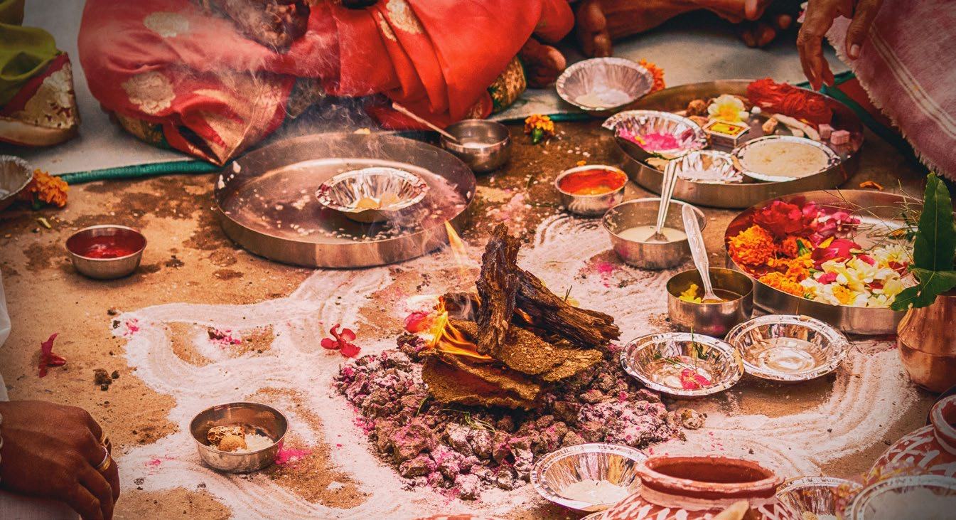 A close-up of a Hindu puja ritual with a small ceremonial fire, flowers, and various offerings arranged on the ground.
