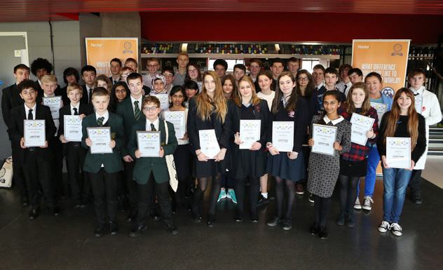 A large group of students posing with certificates at an event, likely an awards ceremony or competition, with banners in the background.