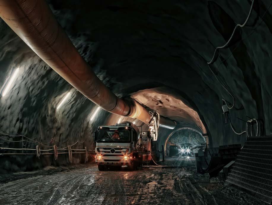 A construction truck with its headlights on drives on a muddy road inside a dark tunnel, with a large ventilation pipe overhead and other construction equipment visible further down.