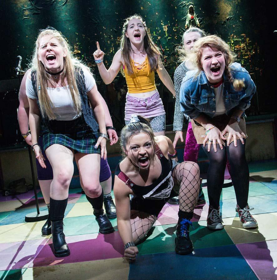 Five young women in punk-rock attire on stage, expressing emotion through singing or shouting, likely a performance.