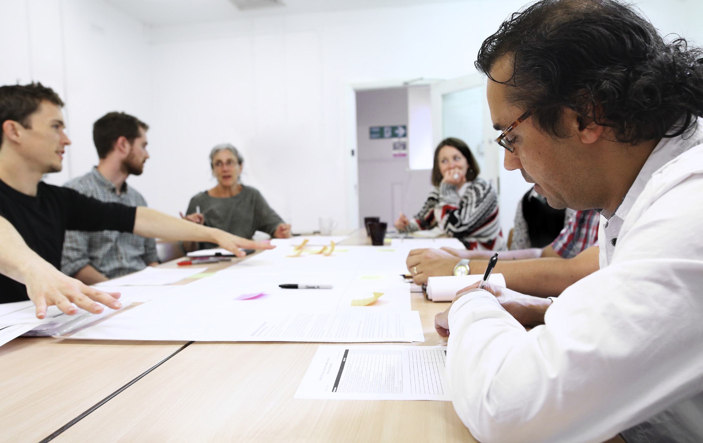 Group of people collaborating around a large table covered with documents and notes, illustrating a workshop or brainstorming session.