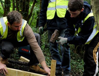 Two men in high-visibility vests working on a wooden structure in a forest, one hammering.