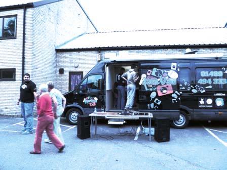 A black "Jamboree" branded van with open side doors and a table with speakers, surrounded by people at an outdoor event.