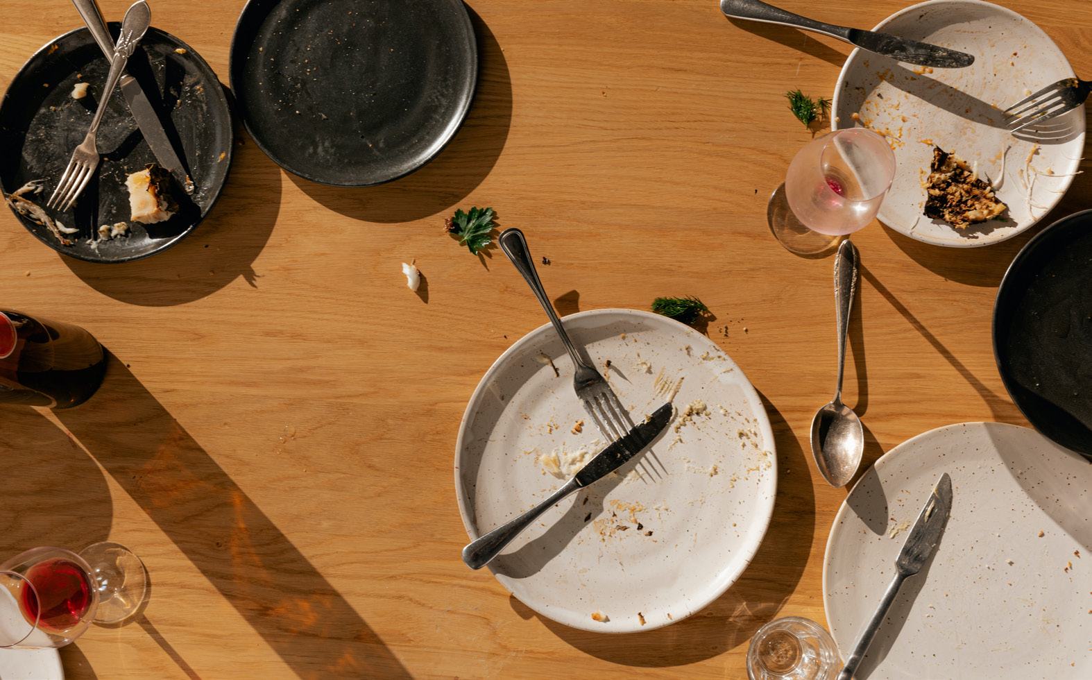 Overhead view of a wooden dining table with several dirty plates, used cutlery, and leftover food scraps after a meal.