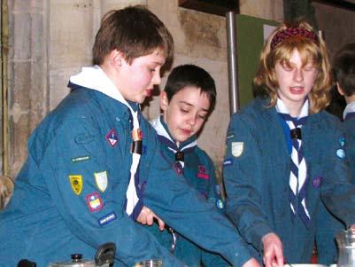 Three children in blue scout-like uniforms with badges and neckerchiefs, engaged in an activity around a table indoors.