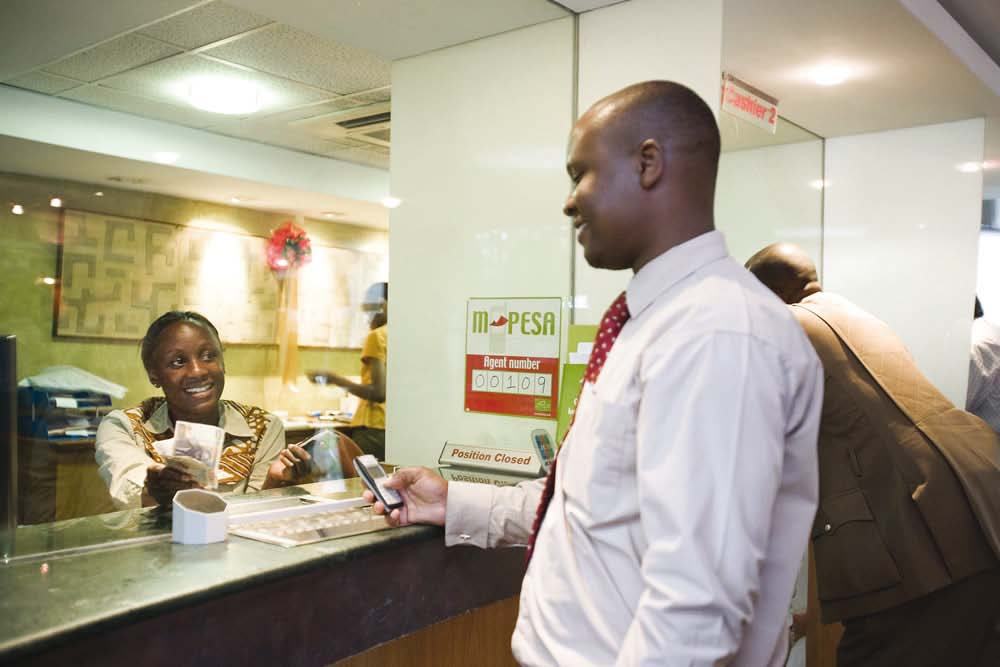 A man uses a mobile phone to conduct a transaction with a female teller behind a glass counter at an M-PESA service point.
