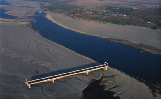 Aerial view of a wide riverbed with a narrow dark blue river channel flowing through sandy banks, crossed by a long, low concrete bridge or causeway.