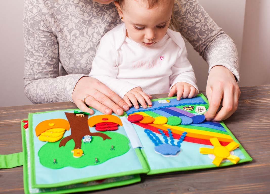 A young child with an adult's hands, interacting with a colorful fabric 'busy book' that features different textures and activities.