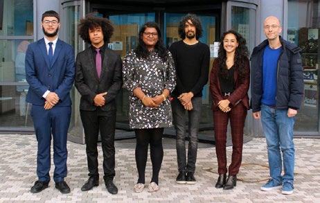 Group photo of six young adults standing side-by-side outdoors in front of a modern building entrance.