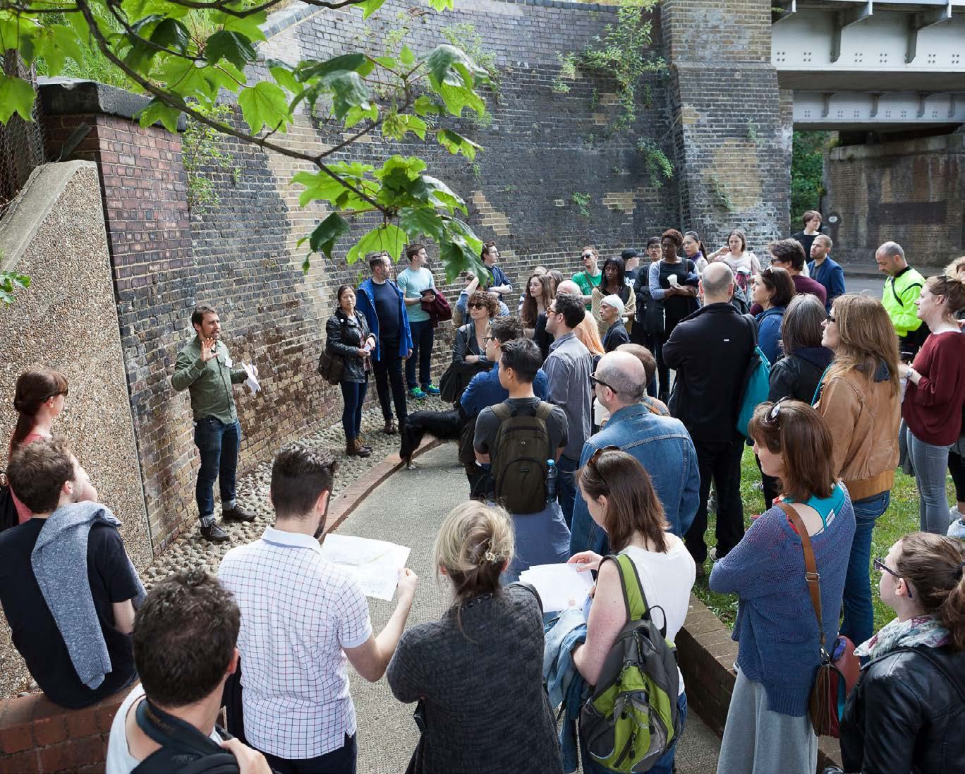A group of people on an outdoor tour, some looking at maps, listening to a speaker next to a brick wall.