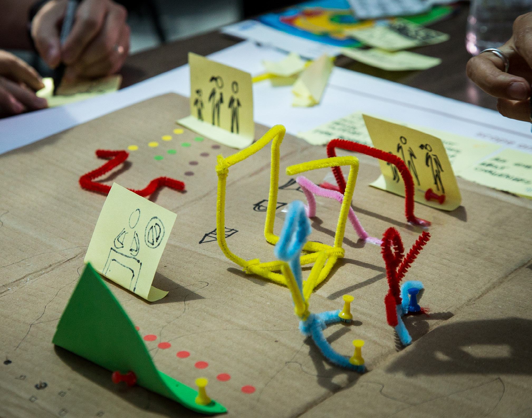 Tabletop view of a workshop with pipe cleaner models, sticky notes with drawings, and pushpins arranged to represent ideas or scenarios.