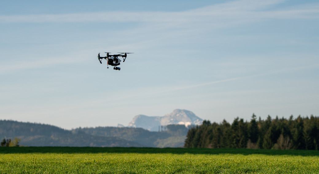 A drone with multiple rotors and a camera flies over a green field with distant mountains under a blue sky.