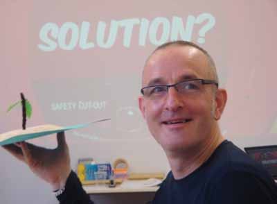 A man smiles while holding a small handmade model of an island with a palm tree, with the word "SOLUTION?" projected on a screen in the background.