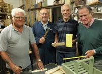 Four older men in a woodworking shop, holding tools and working on a wooden project, smiling.