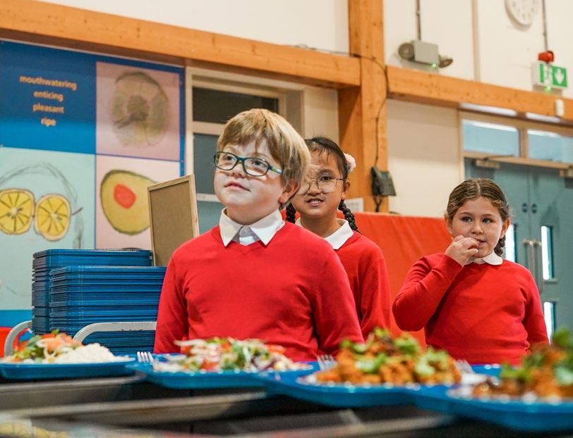 Three elementary school students in red uniforms and white shirts look up, standing in front of prepared meals in a school hall with educational posters on healthy food.