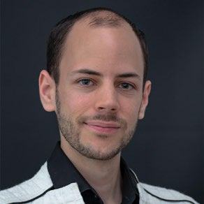 Headshot of a man with short hair and a beard, wearing a black shirt and a white sweater, smiling.
