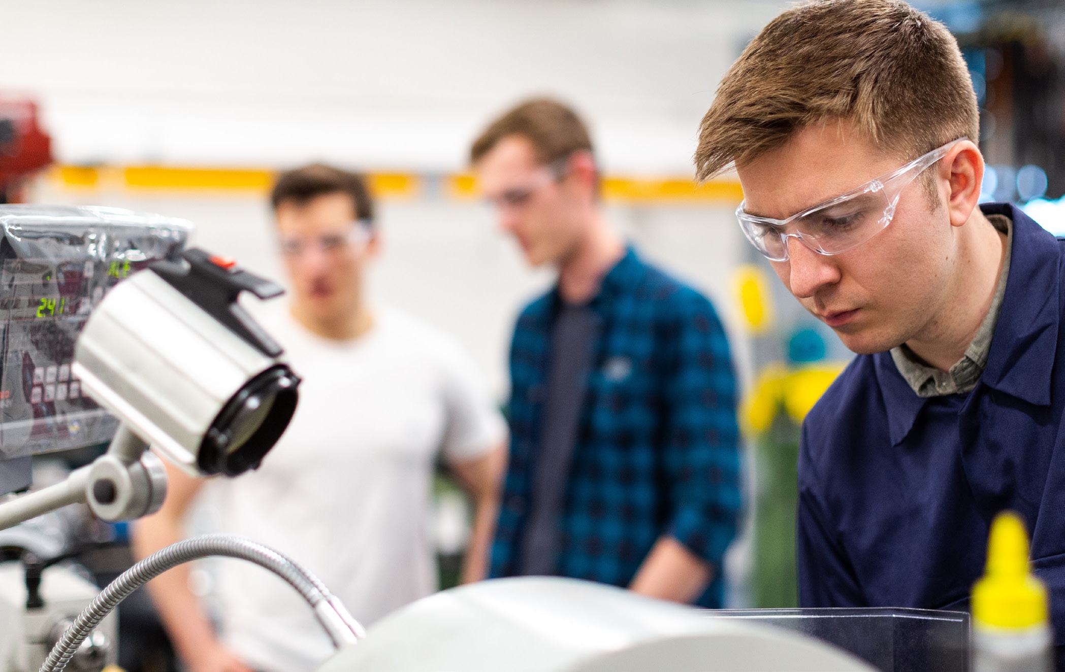 Young man in safety glasses working on machinery in a workshop, with two colleagues observing in the background.