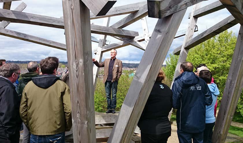 A man speaking to a group of people gathered inside a large, open wooden architectural structure, giving a tour or presentation.
