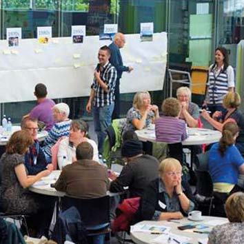 A group of people seated at round tables discussing, with a wall of charts and sticky notes behind them, indicating a workshop or brainstorming session.