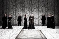 Black and white image of actors in dark robes on a stage, possibly a dramatic scene from a play or opera.