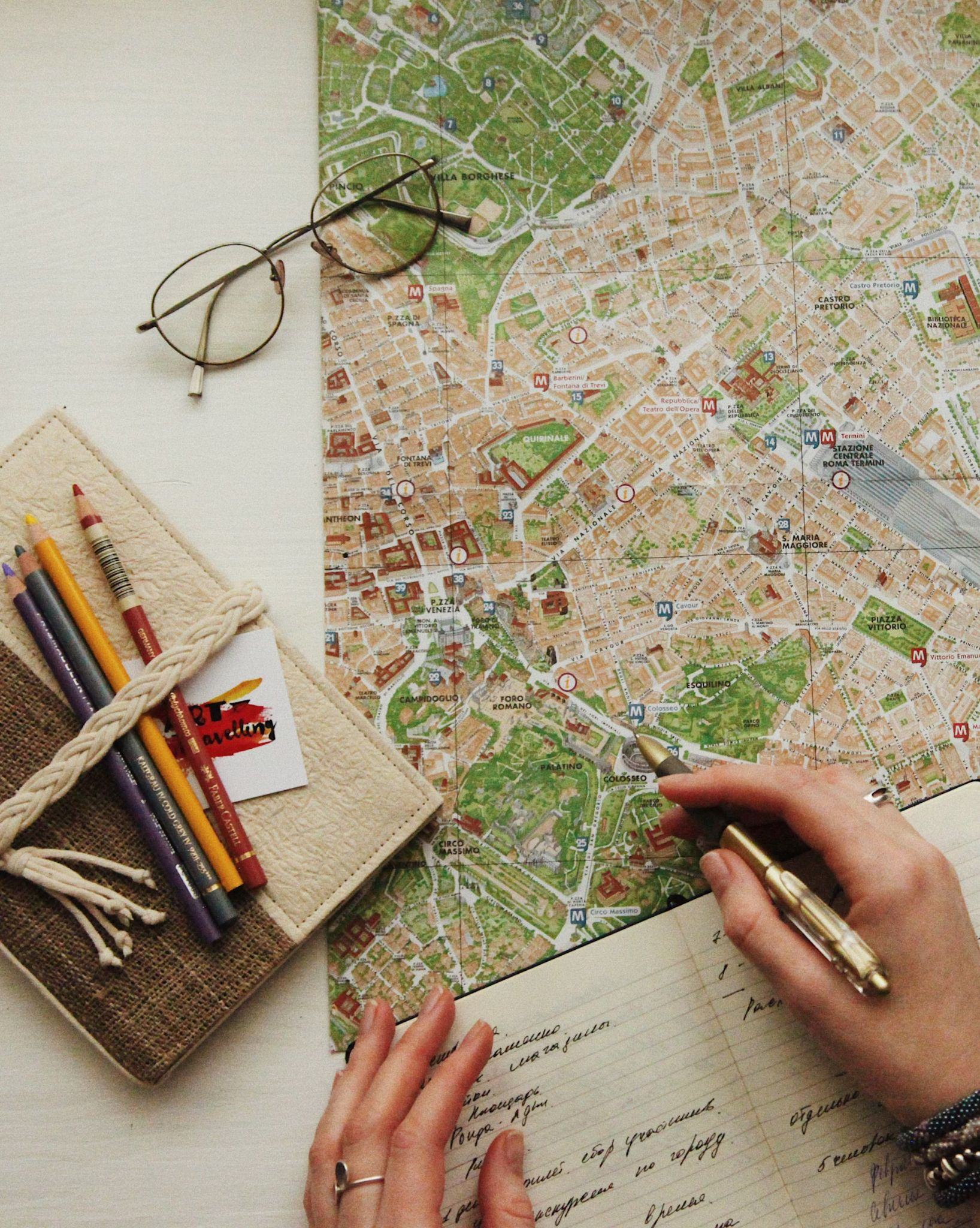 Overhead view of a person using a detailed city map, with glasses, a notebook, and colored pencils on a white table.