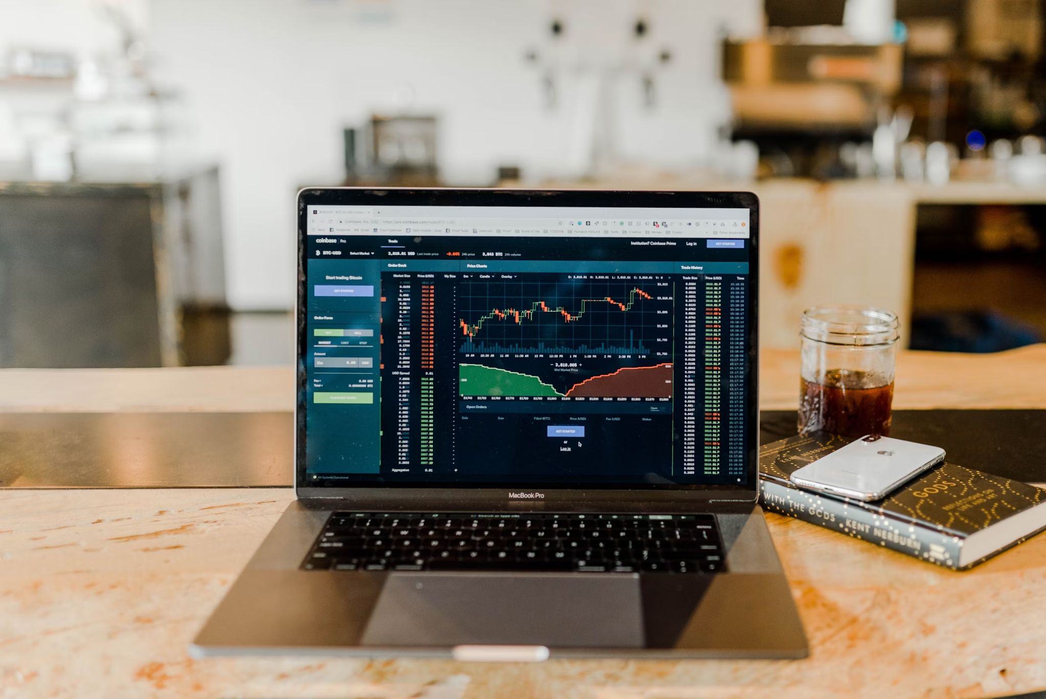 Laptop screen displaying cryptocurrency trading charts and data, with a phone, notebook, and a glass of iced coffee nearby on a table.